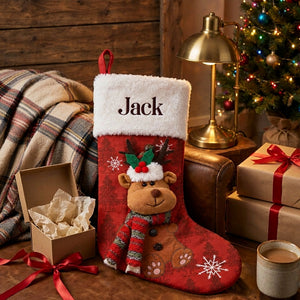 Personalized red stocking with a plush reindeer beside an open kraft gift box lined with ivory tissue paper