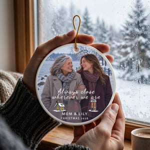 Two hands holding the personalized long distance ornament for Mom up by its gold cord in a bright room